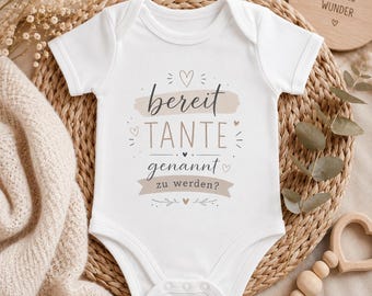 May include: A white short-sleeved baby bodysuit featuring the German text 'bereit TANTE genannt zu werden?' printed in dark gray and beige. The design includes decorative heart icons and brush stroke accents, with a snap-button closure at the bottom.