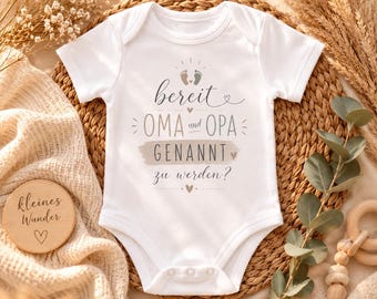 May include: A white short-sleeved baby bodysuit featuring the text "bereit OMA und OPA GENANNT zu werden?" in dark gray and beige, accompanied by small heart icons and a pair of baby footprints printed on the chest area.