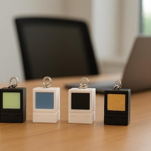 Peut inclure: Quatre porte-clés miniatures en forme d'ordinateur avec des écrans de différentes couleurs : vert, bleu, noir et or. Les porte-clés sont noirs et blancs, avec un anneau argenté en haut. Ils sont disposés sur une surface en bois brun clair.