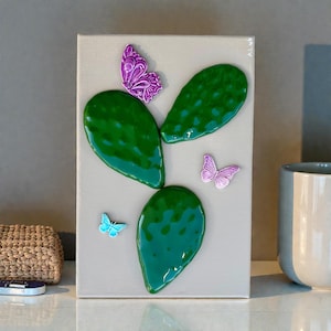 Könnte beinhalten: Ein Leinwandkunstwerk mit einem grünen Kaktusdesign mit dreidimensionalen Elementen und mehreren bunten Schmetterlingen. Die Schmetterlinge sind in Lila- und Blautönen gehalten und verleihen dem Kunstwerk eine verspielte Note. Der Hintergrund ist ein neutrales Grau.