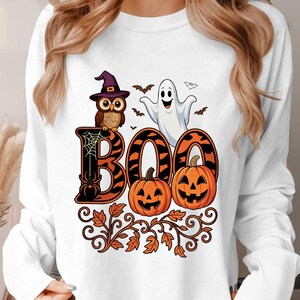 May include: A white long-sleeve sweatshirt featuring a graphic print of the word BOO in orange and black. The design includes a brown owl wearing a purple hat, a white ghost, two orange jack-o'-lanterns, a spider, and orange autumn leaves.