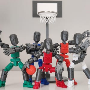 May include: Five gray articulated action figures playing basketball. The figures are wearing different colored jerseys with the number 23 on them. The figures are standing on a gray base with a basketball hoop above them.