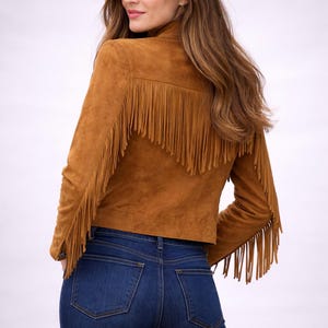 Puede incluir: Chaqueta de ante marrón con flecos en la espalda y las mangas. La chaqueta se combina con vaqueros de mezclilla azul oscuro. La modelo está de espaldas, con el pelo largo castaño.