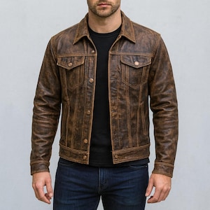 Könnte beinhalten: Braune Lederjacke mit Used-Look. Die Jacke hat zwei Brusttaschen mit Knopfverschluss, eine Knopfleiste und einen Kragen. Das Model trägt ein schwarzes T-Shirt und blaue Jeans.