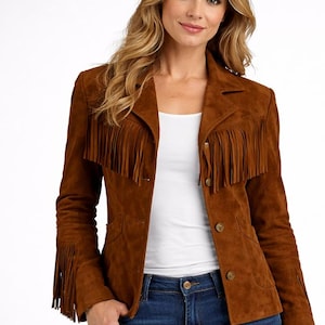 Puede incluir: Chaqueta de ante marrón con flecos en las mangas y el pecho. La chaqueta tiene un cuello clásico, cierre de botones y dos bolsillos delanteros. La modelo lleva vaqueros azules y una camiseta blanca.