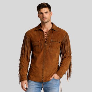 Può includere: Una camicia in pelle scamosciata marrone con maniche lunghe e frange sulle maniche. La camicia ha un davanti allacciato e due tasche sul petto. Il modello indossa jeans blu.