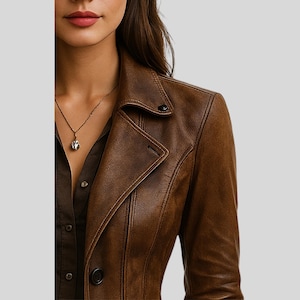 May include: A brown leather jacket with a notched collar and button closure. The jacket has a tailored fit and visible stitching details. A gold necklace with a pendant is visible. The jacket is a classic outerwear piece.