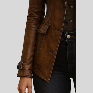 May include: A brown leather jacket with a classic design. The jacket features a notched collar, a button closure, and a small pocket on the chest. The sleeves have a buckle detail at the cuffs. The jacket is paired with dark denim jeans.