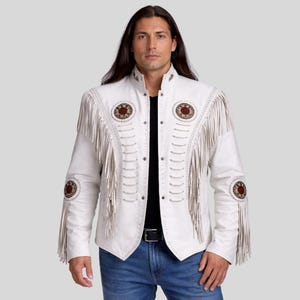 Pode incluir: Casaco de couro branco com detalhes de franjas e patches circulares decorativos. O casaco apresenta gola alta, fecho de botões e franjas marrons. É usado com jeans azuis e uma camisa preta.
