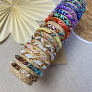 Könnte beinhalten: Eine Sammlung von zwölf zylindrischen Perlenarmbändern, die auf einem beigen Stoffständer angeordnet sind. Jedes Armband weist ein einzigartiges Muster aus kleinen, bunten Glasperlen auf, darunter Blau-, Lila-, Orange-, Gelb-, Weiß- und Brauntöne.
