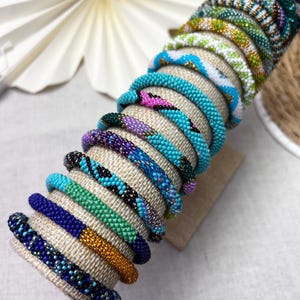 Könnte beinhalten: Eine Sammlung von zwölf zylindrischen Perlenarmbändern, die auf einem strukturierten Ständer gestapelt sind. Jedes Armband weist ein einzigartiges Muster aus kleinen, bunten Glasperlen in Blau-, Grün-, Lila-, Rosa-, Gold- und Weißtönen auf.