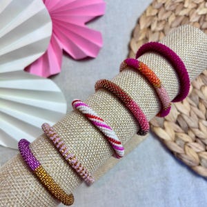 Könnte beinhalten: Eine Sammlung von sechs kreisförmigen Perlenarmbändern, die auf einem zylindrischen Ständer angeordnet sind. Die Armbänder weisen verschiedene Farbkombinationen auf, darunter einfarbiges Magenta, Orange und Pink, Dunkelrot, eine weiß-rote Spirale, Hellrosa sowie Violett mit Gold.