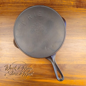 Puede incluir: Una sartén de hierro fundido de color gris oscuro con un mango largo y una superficie de cocción circular. La palabra "ERIE" está grabada en la tapa. La sartén está sobre una superficie de madera. El texto "Work it May, Shine it Must" también es visible.