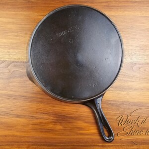 May include: A black cast iron skillet with a long handle, resting on a wooden surface. The word "SIDNEY" is visible on the skillet's base, along with the letters "TN". The skillet is a kitchenware item.