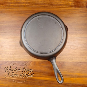 May include: A cast iron skillet with a single handle, viewed from the bottom. The number "5" is embossed on the base. The text "Work it May, Shine it Must" is etched on the wooden surface.