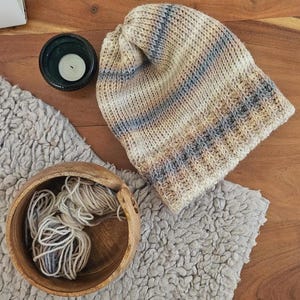 May include: A knitted beanie in beige, blue, and brown tones, placed on a grey, textured surface. A wooden bowl filled with yarn and a small candle in a dark green holder are also visible, suggesting a handmade theme.