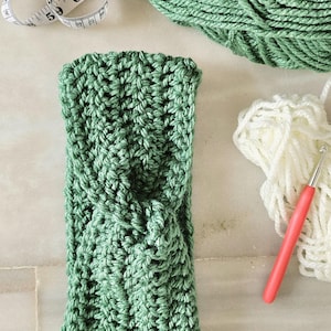 Puede incluir: Una diadema de ganchillo verde hecha a mano con un diseño retorcido. La diadema se muestra con una madeja de hilo verde, hilo blanco, un ganchillo y una cinta métrica.