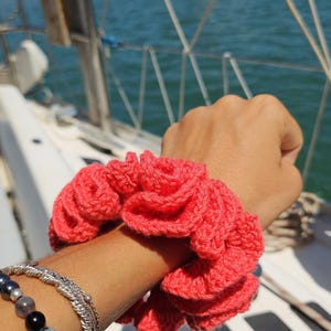 Puede incluir: Una scrunchie de ganchillo de color coral, usada en la muñeca. La scrunchie tiene una textura con volantes y se combina con pulseras de plata y una pulsera de cuentas. El fondo muestra una masa de agua y barcos.