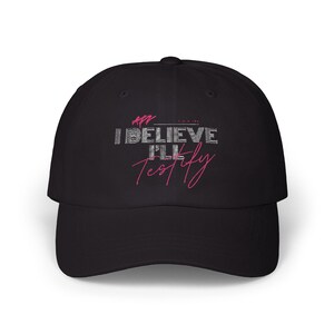 May include: Black baseball cap with white text that reads "I BELIEVE I'LL TESTIFY" in a bold, blocky font. The text "I'LL" is in pink.