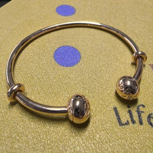 Può includere: Un braccialetto a cerchio color oro con due estremità sferiche decorative. Il braccialetto ha una finitura liscia e lucida e presenta dettagli intricati sugli accenti sferici. Il braccialetto è esposto su una superficie strutturata verde oliva.