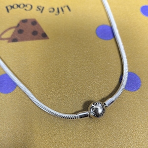 May include: A silver snake chain necklace with a round clasp. The clasp has the letters "OP" and "A" engraved on it. The necklace is on a gold-coloured surface with the words "Life is Good" visible in the background.