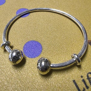 May include: A silver bangle bracelet featuring two spherical charms and two decorative knots. The charms are engraved with the word "PANDORA". The bracelet is presented on a gold surface with purple polka dots.