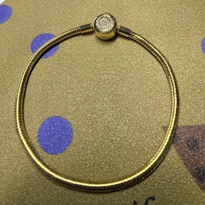 Puede incluir: Una pulsera dorada con un diseño de cadena de serpiente. La pulsera tiene un cierre redondo con un elemento decorativo. La pulsera se muestra sobre una superficie dorada con lunares azules.