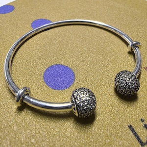 May include: A silver-toned bangle bracelet with two spherical, textured charms. The charms are covered in small, dark-colored stones. The bracelet is resting on a gold-colored surface with purple polka dots.