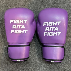May include: A pair of purple boxing gloves with the words "FIGHT RITA FIGHT" printed in white. The gloves have a black wrist strap and are labeled "16-OZ".