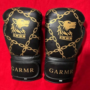 May include: Pair of black boxing gloves with gold chain and wolf head designs. The gloves have the word "GARMR" in gold lettering on the wrist straps. The gloves are set against a red background.