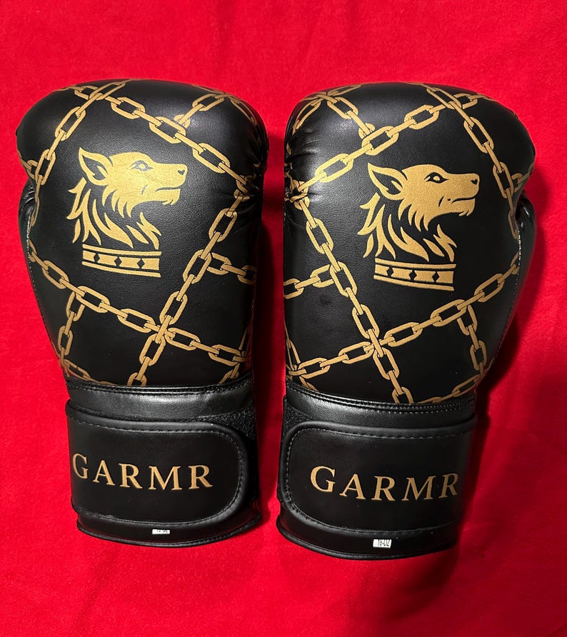 May include: Pair of black boxing gloves with gold chain and wolf head designs. The gloves have the word "GARMR" in gold lettering on the wrist straps. The gloves are set against a red background.