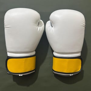 May include: A pair of white boxing gloves with yellow wrist straps. The gloves are made of a smooth material and have a classic design. The wrist straps are accented with black trim and the gloves are shown against a dark green background.