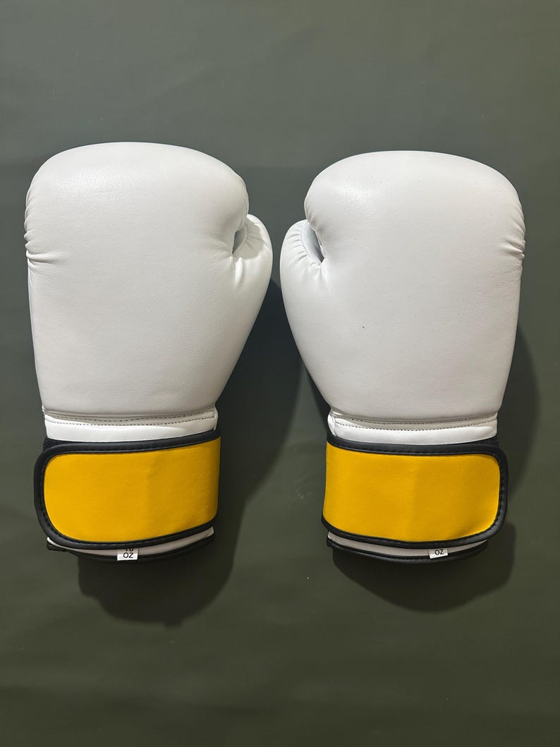 May include: A pair of white boxing gloves with yellow wrist straps. The gloves are made of a smooth material and have a classic design. The wrist straps are accented with black trim and the gloves are shown against a dark green background.