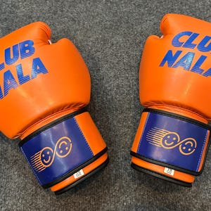 May include: Pair of orange boxing gloves with the text "CLUB NALA" in blue. The wrist straps are blue with a design of two smiling faces. The gloves are 10 oz.