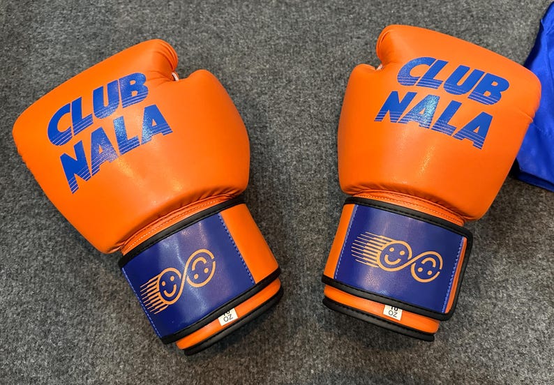 May include: Pair of orange boxing gloves with the text "CLUB NALA" in blue. The wrist straps are blue with a design of two smiling faces. The gloves are 10 oz.