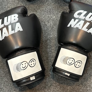May include: Pair of black boxing gloves with the text "CLUB NALA" in white on the top. Each glove has a white patch with two smiling face graphics. The gloves are labeled "10 OZ".