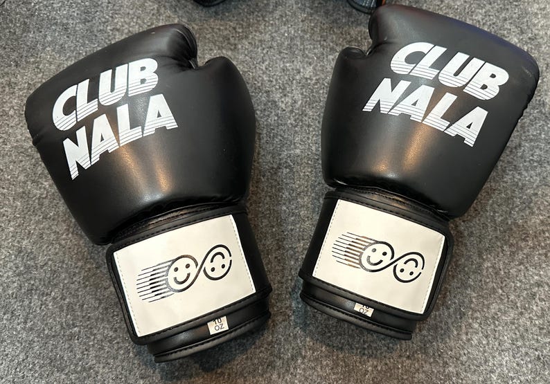 May include: Pair of black boxing gloves with the text "CLUB NALA" in white on the top. Each glove has a white patch with two smiling face graphics. The gloves are labeled "10 OZ".