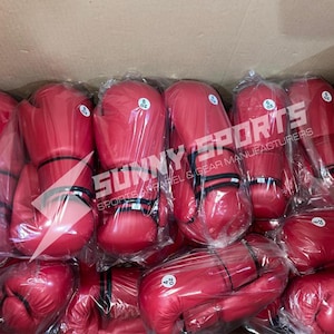 May include: A collection of red boxing gloves, some labeled 8 oz and 4 oz, are packaged in clear plastic. The gloves are stacked inside a cardboard box. The text "SUNNY SPORTS SPORTS APPAREL & GEAR MANUFACTURERS" is visible.