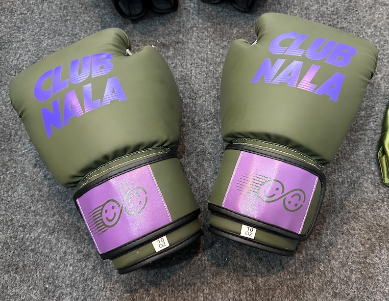 May include: Olive green boxing gloves with the text "CLUB NALA" in purple. The wrist straps are purple with a smiley face design and the text "10 OZ".