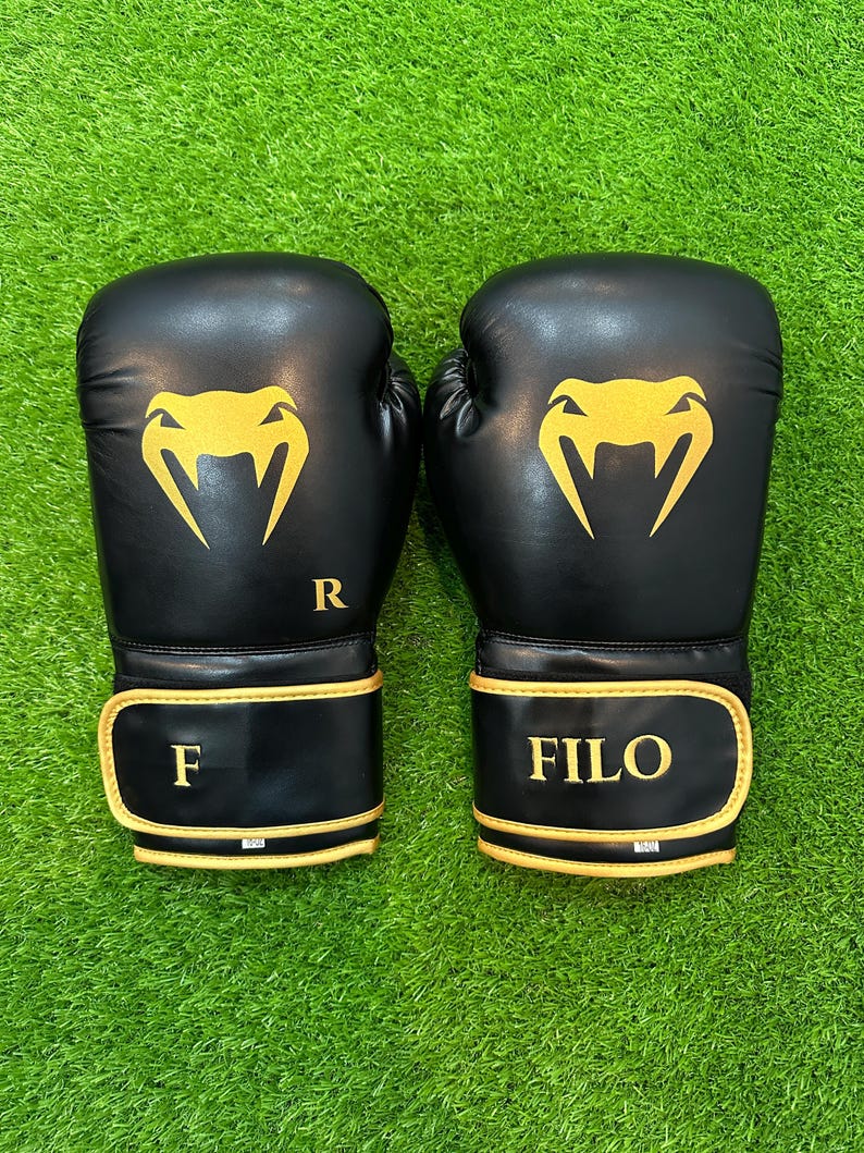 May include: A pair of black boxing gloves with gold accents. Each glove features a gold logo and the letters "R" and "F" on one glove and "FILO" on the other. The gloves are set against a green background.