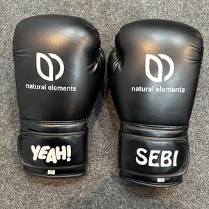May include: A pair of black boxing gloves with white text and logo. The gloves have the words "YEAH!" and "SEBI" on the wrist straps. The logo is a stylized "D" shape with the words "natural elements" below it.