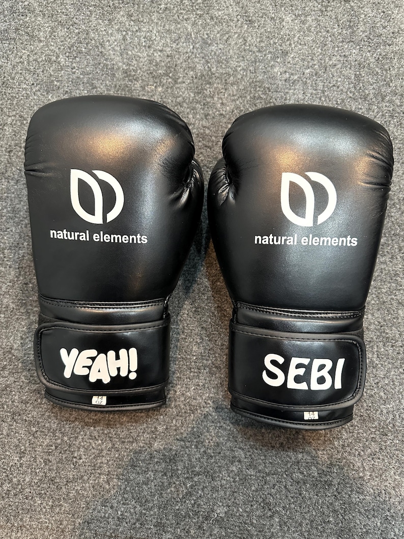 May include: A pair of black boxing gloves with white text and logo. The gloves have the words "YEAH!" and "SEBI" on the wrist straps. The logo is a stylized "D" shape with the words "natural elements" below it.