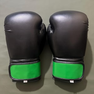 May include: A pair of black boxing gloves with bright green wrist straps. The gloves are made of a smooth material and appear to be new. The wrist straps have a velcro closure and a small white tag with text.