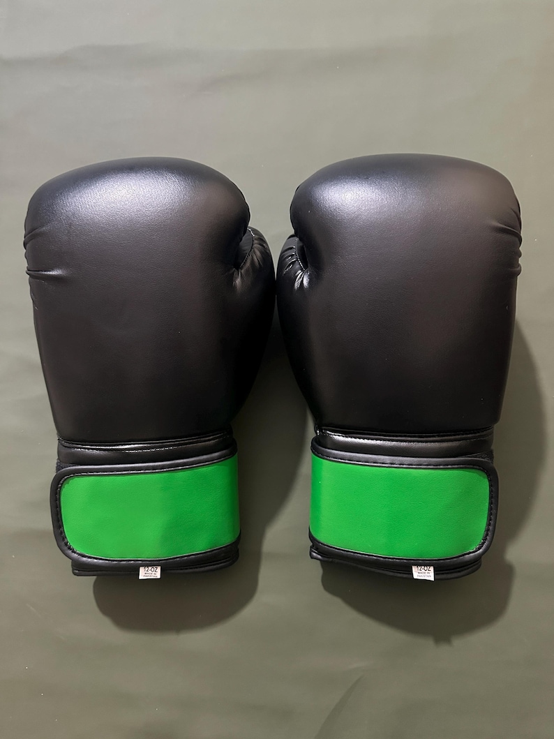 May include: A pair of black boxing gloves with bright green wrist straps. The gloves are made of a smooth material and appear to be new. The wrist straps have a velcro closure and a small white tag with text.