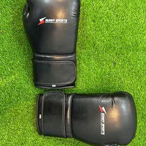 May include: A pair of black boxing gloves with the text "SUNNY SPORTS" printed in white and red. The gloves are made of a smooth material and are set against a green grass background.