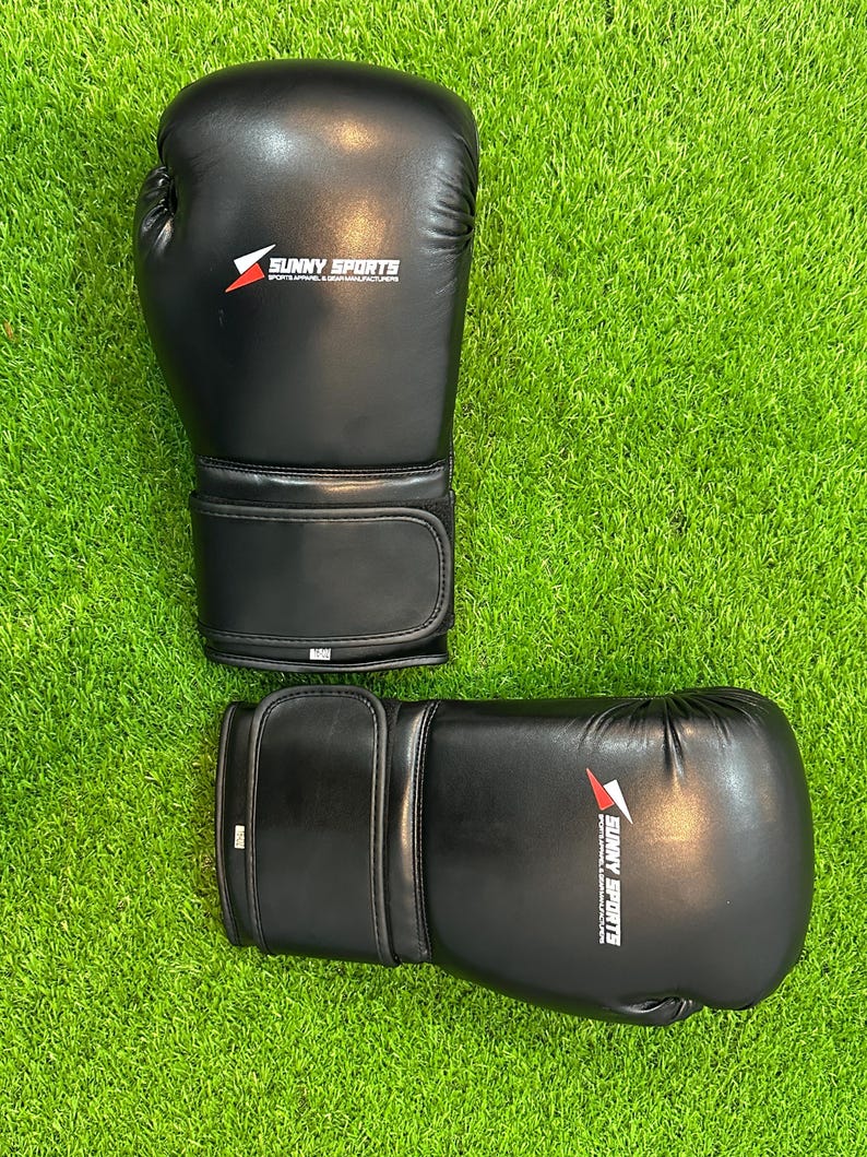 May include: A pair of black boxing gloves with the text "SUNNY SPORTS" printed in white and red. The gloves are made of a smooth material and are set against a green grass background.