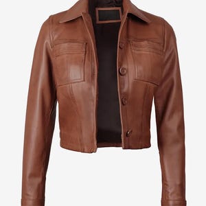 Könnte beinhalten: Braune Lederjacke mit Kragen, zwei Vordertaschen und Knopfverschlüssen. Die Jacke hat lange Ärmel mit geknöpften Manschetten. Das Innenfutter ist schwarz. Die Jacke ist am Bund verkürzt.