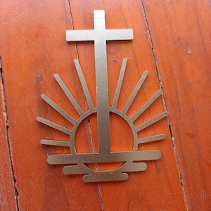 May include: A gold-colored metal religious symbol featuring a cross above a stylized sunburst design. The cross is centered above a circular shape with radiating lines, creating a symmetrical and symbolic image. The symbol is placed on a wooden surface.