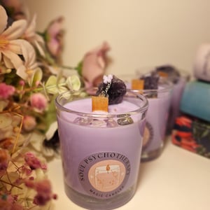 May include: Three lavender-colored candles in clear glass containers, each topped with crystals and a wooden wick. The candles have a label that reads "SOUL PSYCHOTHERAPY MAGIC CANDLES". Flowers are in the background.