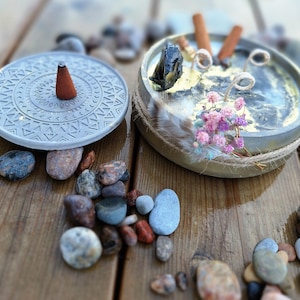 May include: A grey concrete incense holder with a geometric pattern and a brown incense cone. A decorative tray with a crystal, feathers, dried flowers, and incense sticks is next to it. Small stones are scattered around the holders.